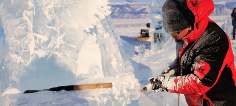 Man in red jacket chisels a block of ice into a sculpture outdoors in winter.