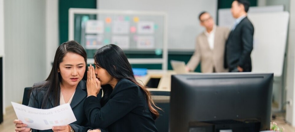 Two colleagues whisper over documents at a busy office desk