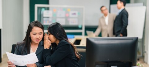Two colleagues whisper over documents at a busy office desk